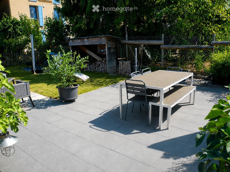 Garden patio with a wooden table, metal chairs, a bench, a wicker chair, and a potted plant. The area is surrounded by greenery and includes a wooden shed and a stack of firewood.