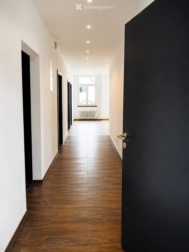Bright, clean hallway with wooden flooring, white walls, and recessed ceiling lighting. Features several doors and a window at the far end.