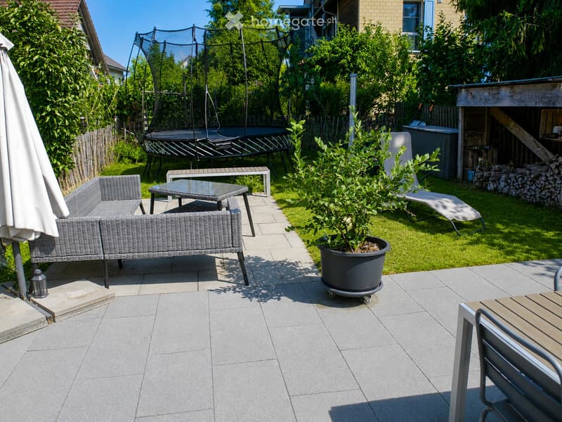 A well-maintained garden with a patio area featuring a grey wicker sofa, a glass-top table, a potted plant, and a sun lounger. A trampoline is visible in the background along with a wooden shed and a stack of firewood.