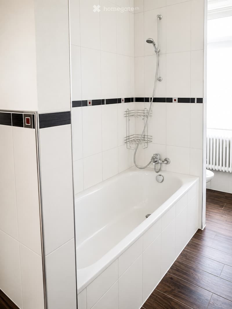 Modern bathroom with a white bathtub, glass shower enclosure, showerhead, and a small shower basket. The walls are tiled in white with black accents, and the floor is wooden.