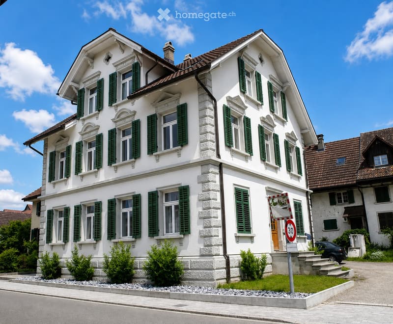A two-story white house with green shutters, multiple windows, and a brown tiled roof. The house has a small garden with shrubs and a paved driveway.