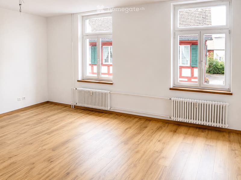 Empty room with light wooden flooring, white walls, and two radiators. Windows offer views of neighboring houses with red facades and green shutters.