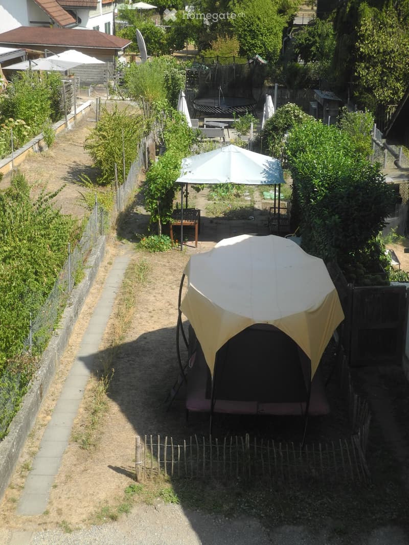 Garden with a trampoline, patio furniture, and umbrellas, surrounded by greenery and a wooden fence.