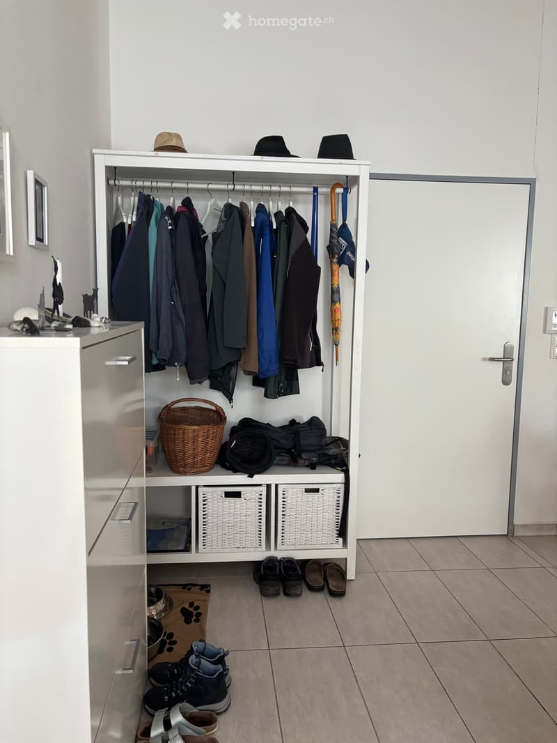 A white wardrobe with a hanging rail, two baskets, and a wicker basket on the lower shelf. A white cabinet with three drawers is on the left side. A few shoes are placed on the floor in front of the cabinet. A white door is on the right side. A few frames