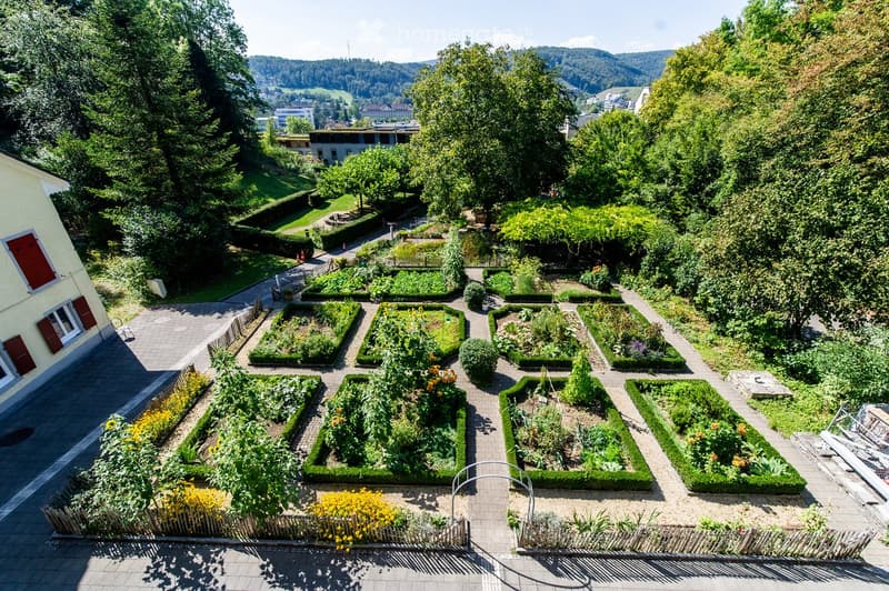 A well-maintained garden with neatly arranged flower beds, pathways, and surrounding greenery, featuring a variety of plants and shrubs.