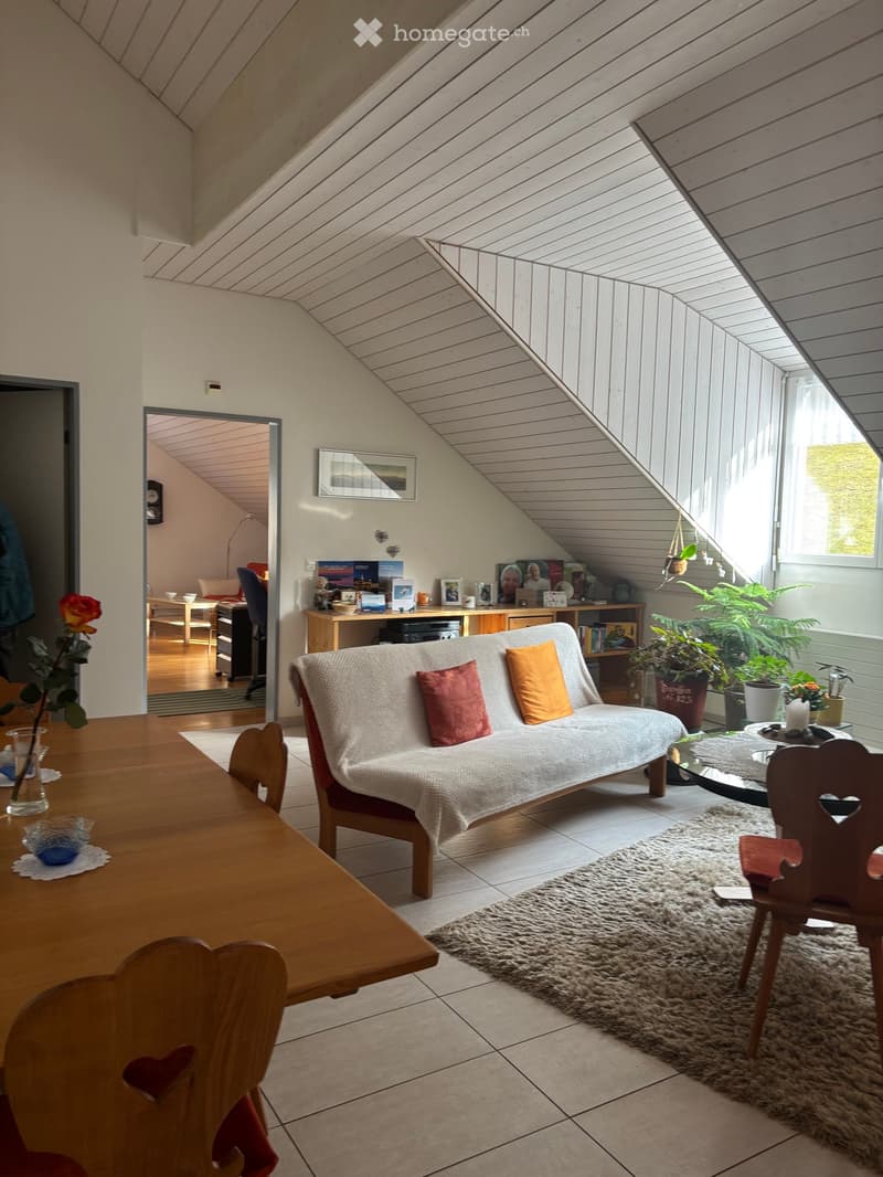 A cozy living room with a sofa, wooden table, chairs, carpet, and plants. The room has a wooden desk, a shelf with books, and a picture frame on the wall. The room has a window with a view of the outside.