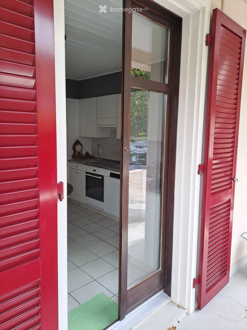 Modern kitchen with white cabinets, stainless steel appliances, and a tiled floor, visible through an open doorway with red shutters.