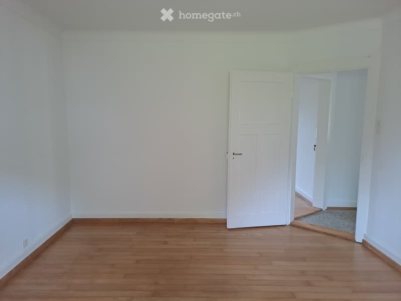 Empty room with light-colored walls and wooden flooring, featuring a closed white door and an open doorway leading to another room.