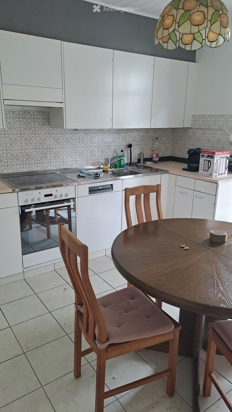 Modern kitchen with white cabinets, a round wooden dining table with two chairs, an oven, dishwasher, and sink area with various appliances and items.