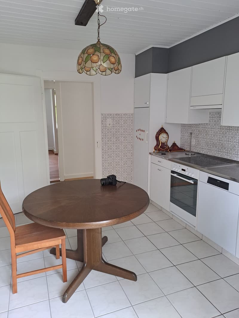 Modern kitchen with white cabinets, a round wooden dining table, a chair, a camera on the table, and a decorative clock on the counter.