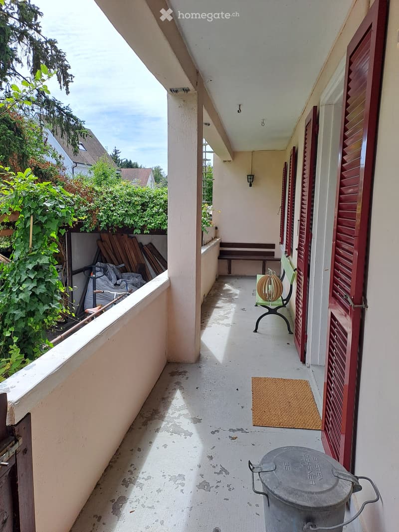 Balcony with a bench, small table and chair, doormat, and trash bin. Red shutters on the right side.