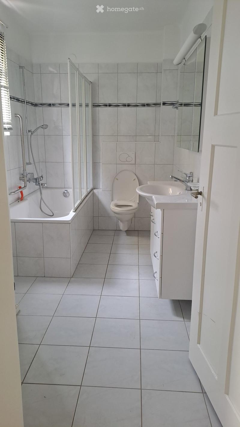 Modern bathroom with white tiles, bathtub, toilet, sink, and mirror. Features a glass-enclosed shower area with a handheld showerhead.