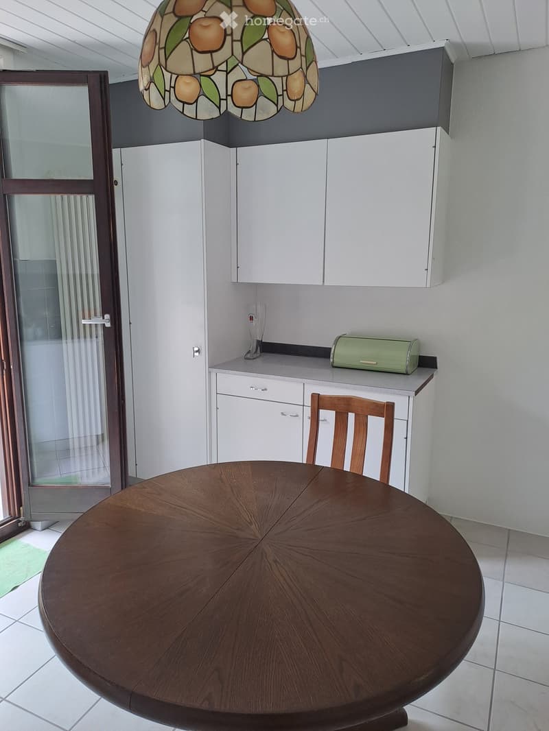 A modern kitchen with white cabinets, a round wooden dining table, a wooden chair, and a decorative stained-glass pendant light.