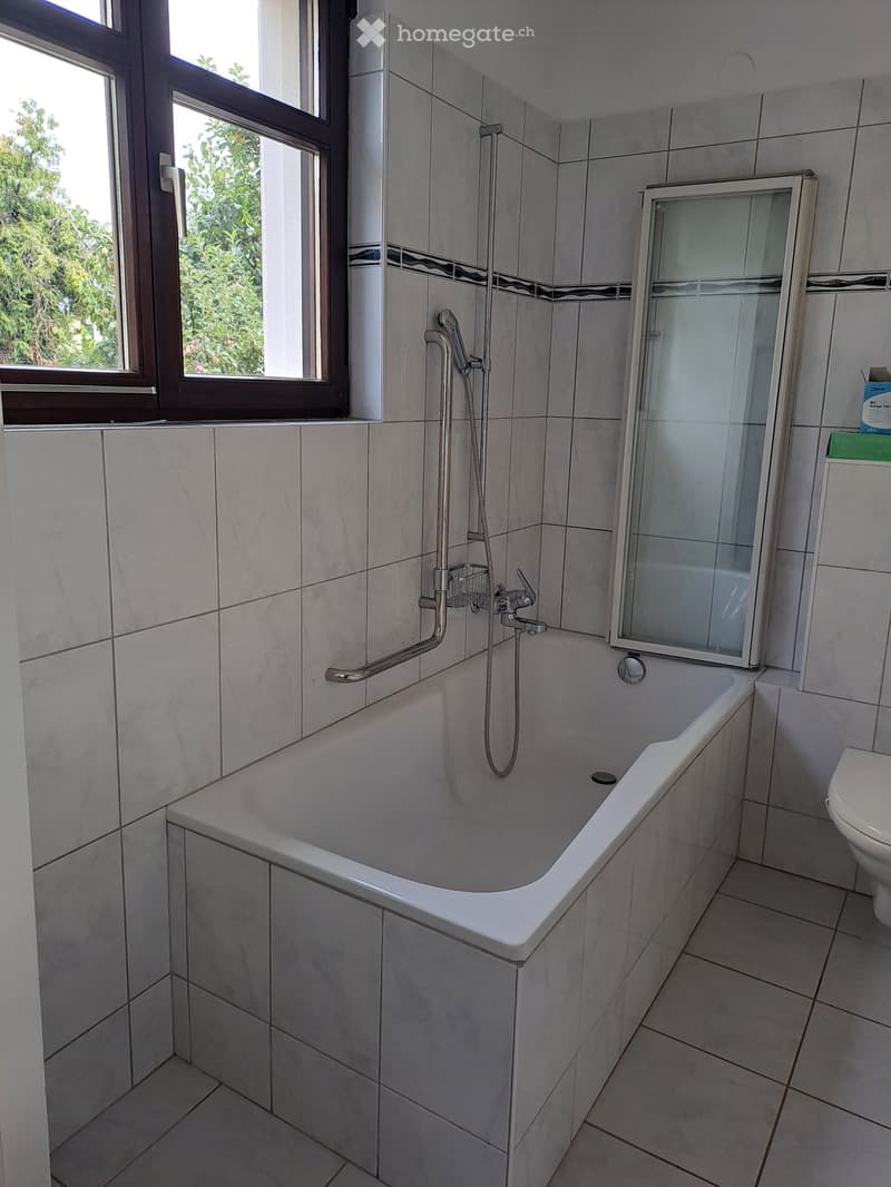 Modern bathroom with a bathtub, shower, and toilet. Features tiled walls and floor, a large window with a view of greenery, and a glass shower enclosure.
