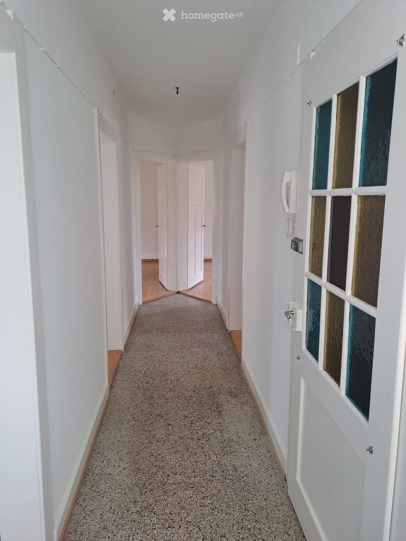 A long, narrow hallway with white walls, carpeted floor, and wooden baseboards. Doors lead to other rooms, and a telephone is mounted on the wall.