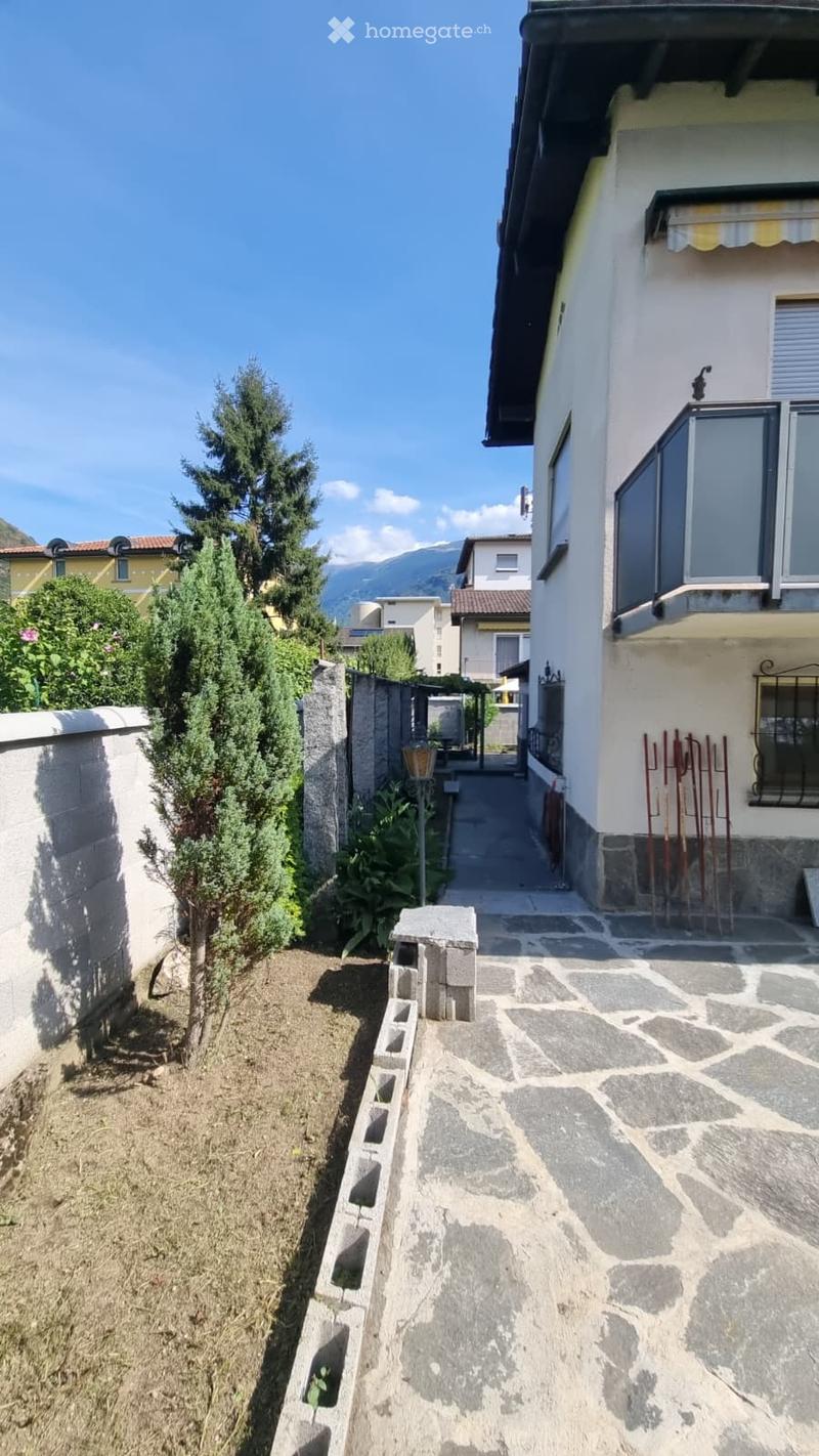 A narrow garden pathway leads to a house with a stone wall on one side and a small tree on the other. The pathway is made of stone slabs, and the house has white walls and dark shutters.