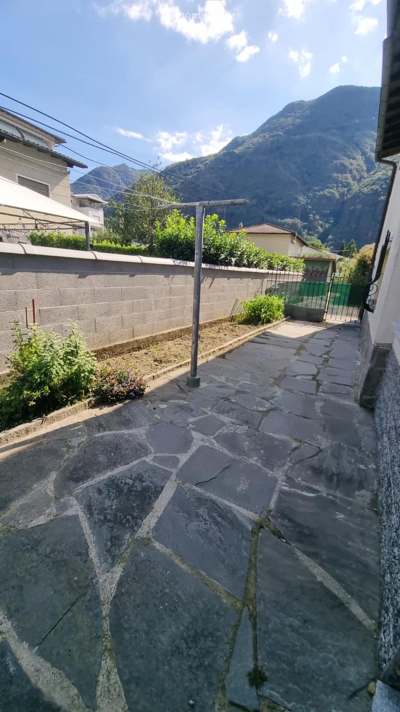 Narrow stone-paved pathway bordered by a low concrete wall, with small patches of greenery and a clothesline pole, leading to a gated area with more greenery and a view of mountains in the background.