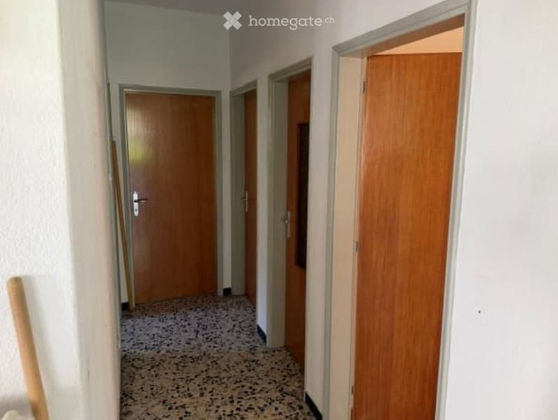 A narrow hallway with wooden doors on both sides, featuring a speckled tile floor and white walls.