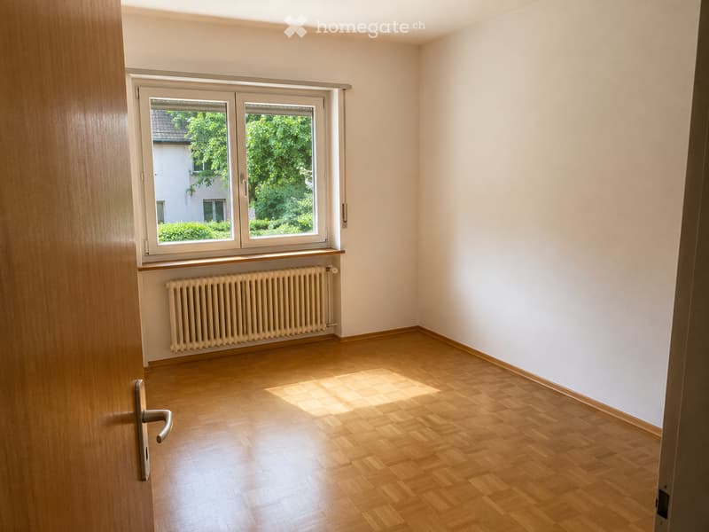 Empty room with wooden flooring, white walls, a window with white frame, and a radiator below the window.