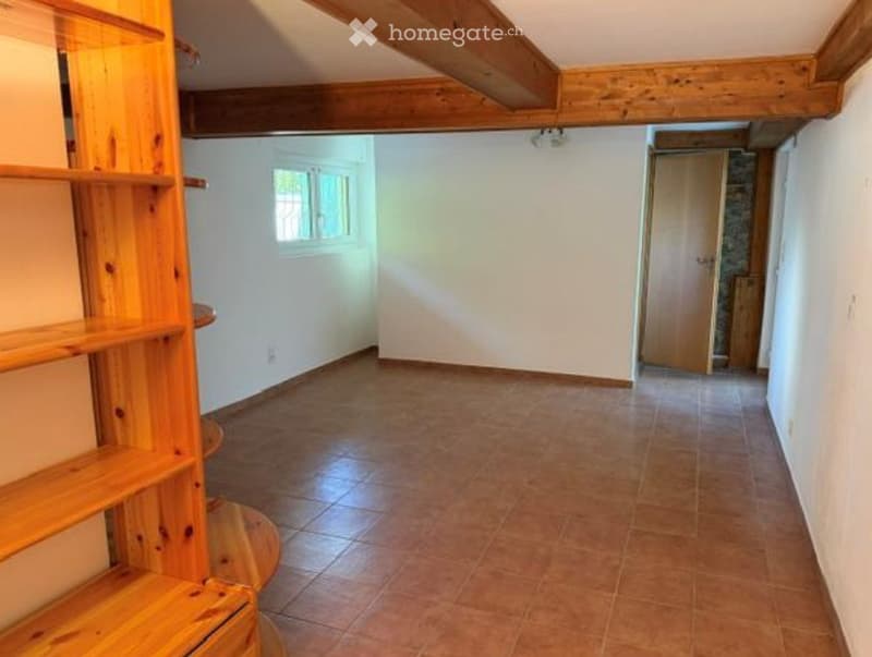 Empty room with wooden beams on the ceiling, tiled floor, and a small window. There is a wooden shelf on the left side and a closed wooden door at the far end.