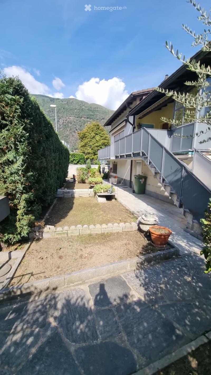Garden area with gravel ground, small plants, a stone pathway, and a house with a balcony in the background.