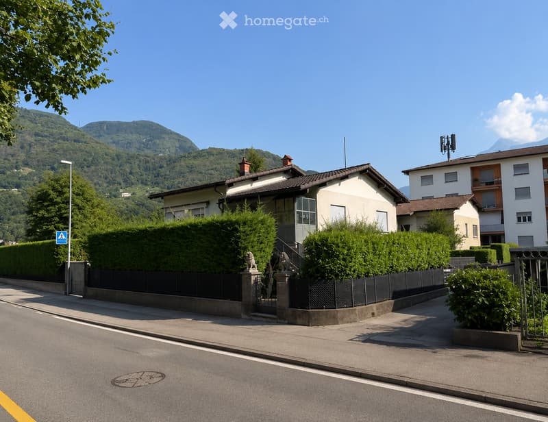 A two-story house with a tiled roof, surrounded by greenery and a black metal fence, with a view of mountains in the background.