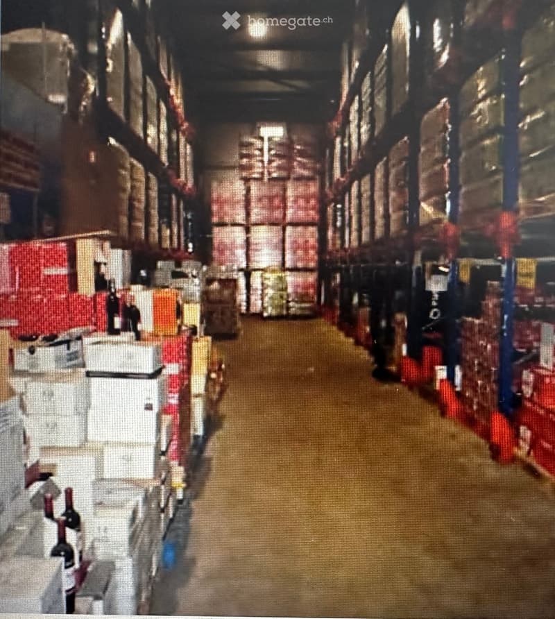 A long, narrow warehouse corridor with metal shelving units on both sides, filled with various boxes and storage items. The floor is concrete, and the ceiling has exposed industrial lighting.
