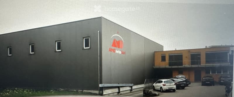 Modern industrial building with a grey facade, logo, and adjacent parking area with several cars.