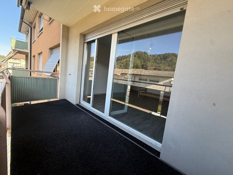Balcony with large glass sliding doors, black carpeted floor, and a view of surrounding buildings and greenery.