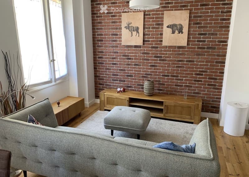Living room with exposed brick wall, grey sofa, ottoman, wooden coffee table, side table, and artwork.