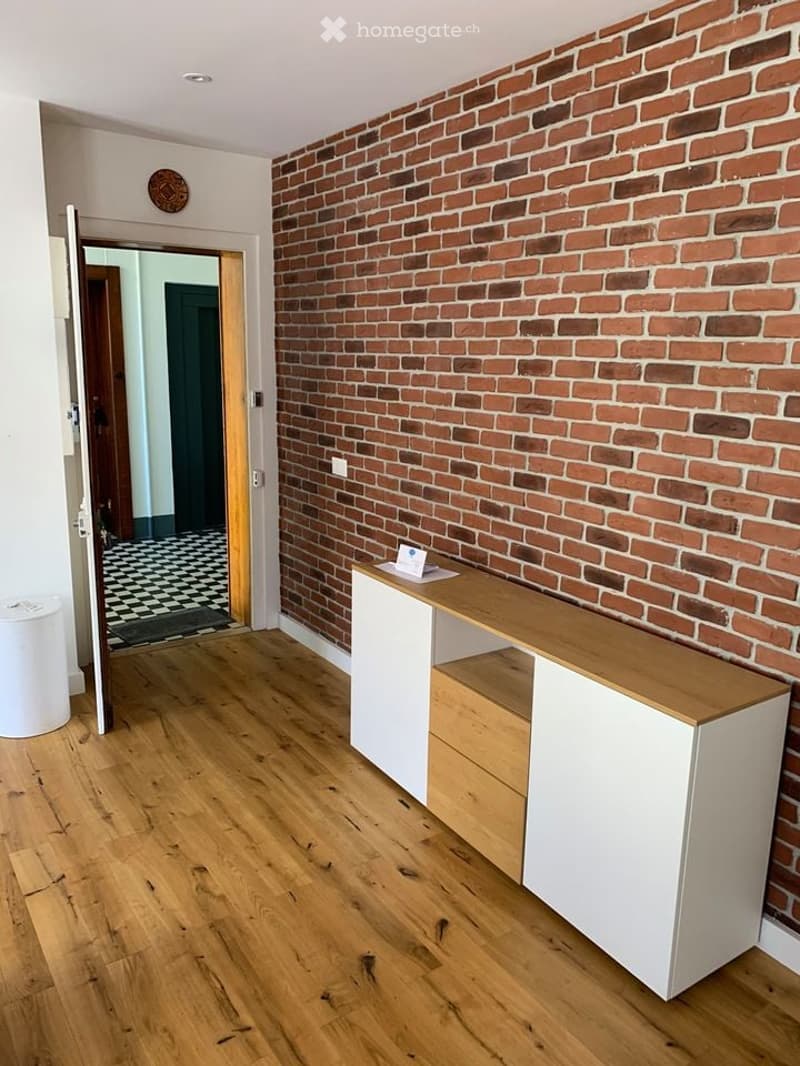 Empty room with exposed brick wall, wooden floor, white cabinet with drawers, and a small table with a card on it. An open doorway leads to another room with checkered flooring.