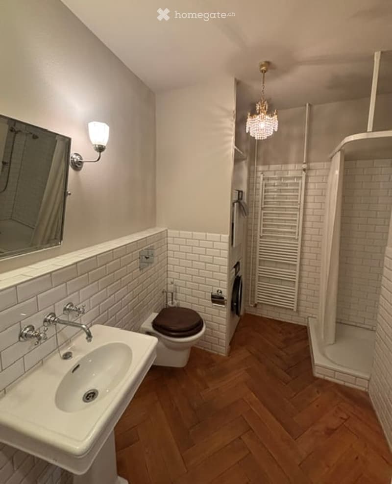 Modern bathroom with white brick tiles, wooden herringbone flooring, a sink with a modern faucet, a toilet, a shower area, and a decorative chandelier light fixture.