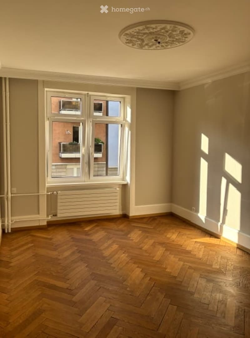 Empty room with wooden herringbone flooring, large window with white frame, radiator below window, and decorative ceiling light fixture.