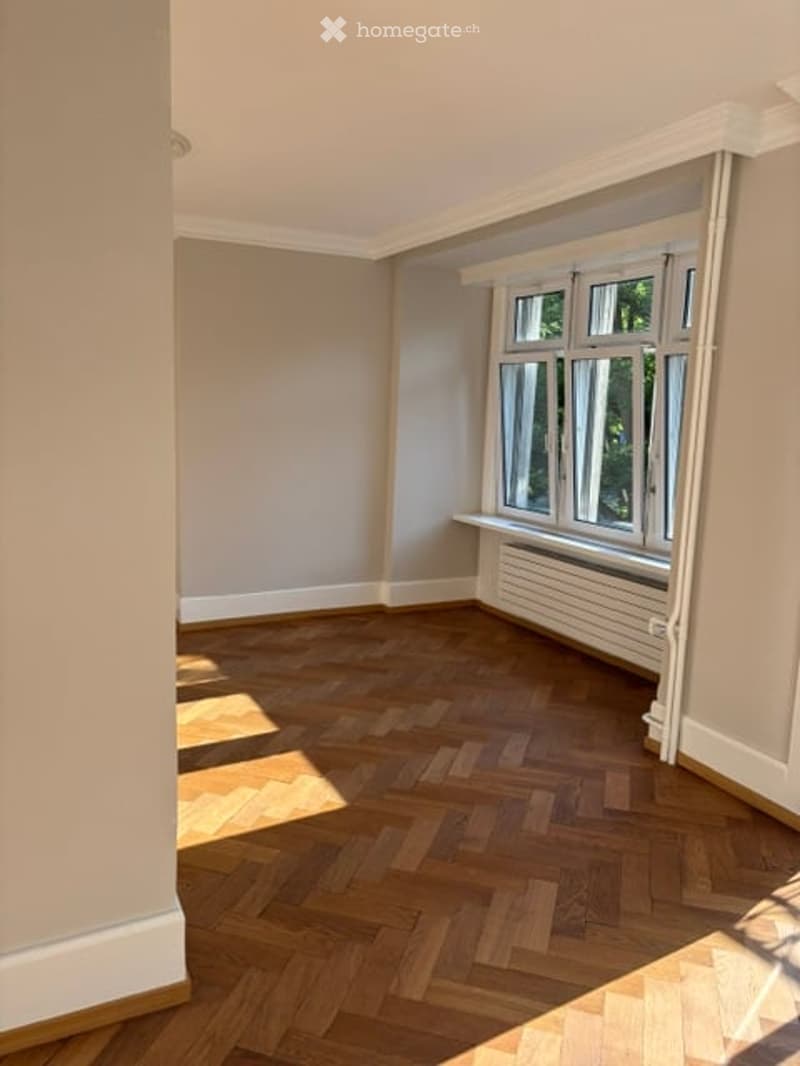 Empty room with wooden herringbone flooring, large windows, and white walls.