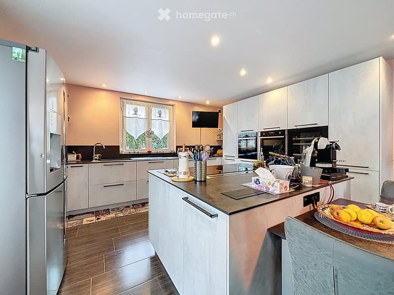 Modern kitchen with white cabinets, stainless steel appliances, and a central island. Features include a refrigerator, oven, coffee maker, sink, and various kitchen utensils. The island has a countertop with a bowl of fruit and a cup.