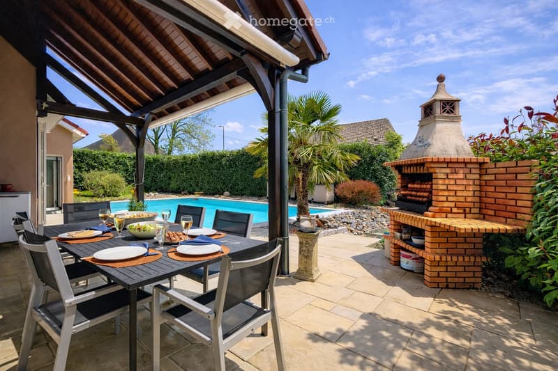 Outdoor patio with a dining table, chairs, and a brick oven. A swimming pool is visible in the background, surrounded by greenery and a covered seating area.