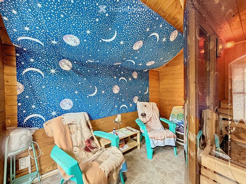 Cozy room with wooden walls, a starry night sky mural, two chairs with blankets, a small table with a candle and lamp, and a view through a glass door.