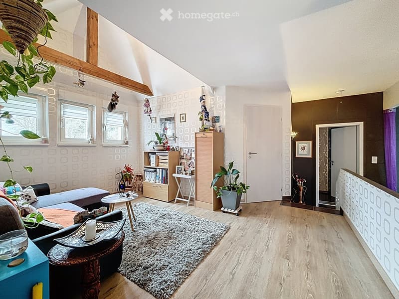 A cozy living room with wooden floors, a patterned rug, a sofa, a coffee table, a bookshelf, and several plants. The room features large windows and a slanted ceiling with exposed wooden beams.