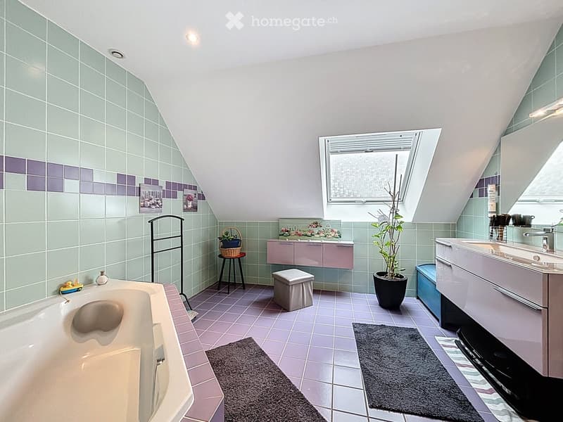 Bathroom with a bathtub, sink, vanity, stool, potted plant, towel rack, and picture frames. The room features light green and purple tiles, a skylight window, and a small window with blinds.
