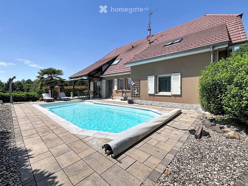 Outdoor area with a swimming pool, surrounded by a tiled patio, lounge chairs, and a house with a tiled roof and shutters.