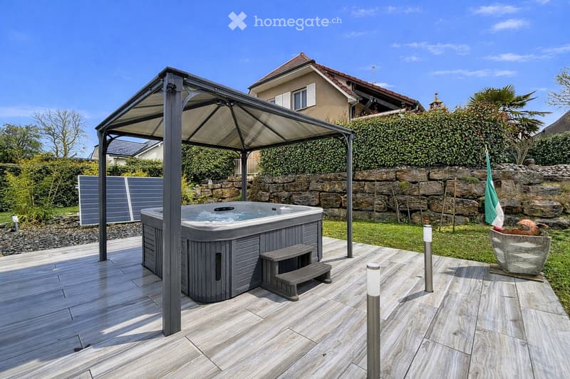 Outdoor patio with a hot tub under a metal canopy, wooden decking, stone wall, and surrounding greenery.