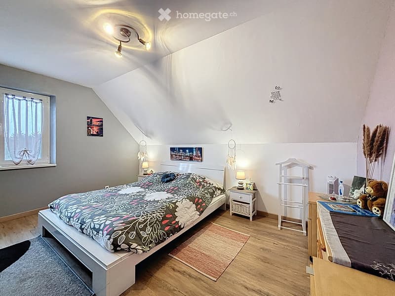 Cozy attic bedroom with wooden flooring, slanted ceiling, and modern decor including a bed, nightstand, lamp, and decorative items.