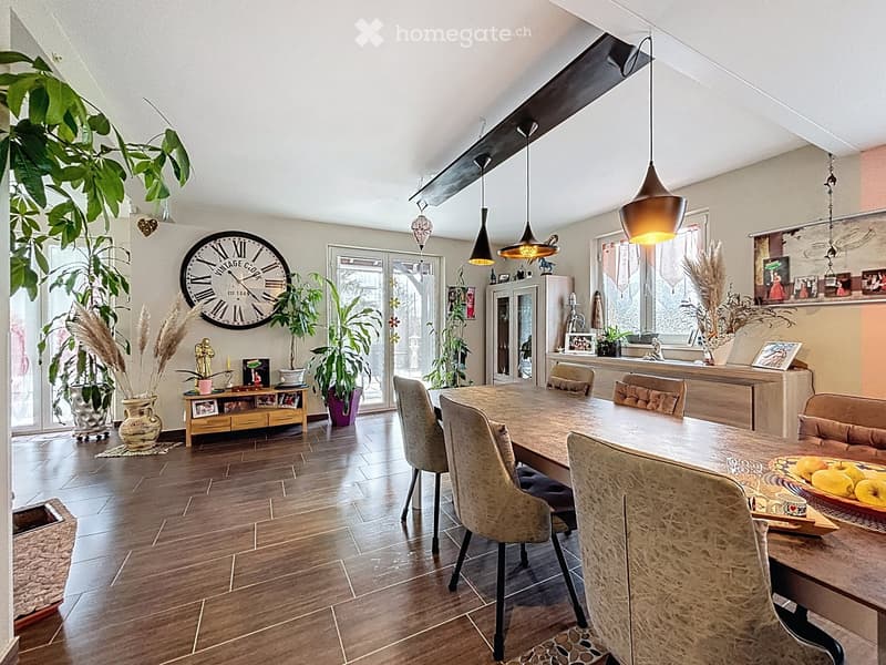 Dining room with wooden floor, large dining table with chairs, pendant lights, decorative clock, cabinet, vases, and potted plants.
