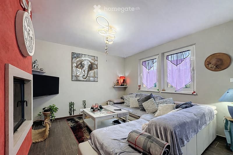 A modern living room with a grey sectional sofa, coffee table, TV, decorative clock, artwork, and plants. The room features large windows with purple curtains and a stylish wall-mounted light fixture.