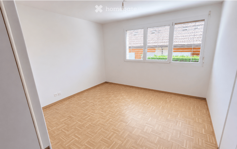 Empty room with light wooden herringbone flooring and white walls. Large window with blinds offers a view of a neighboring building and greenery.