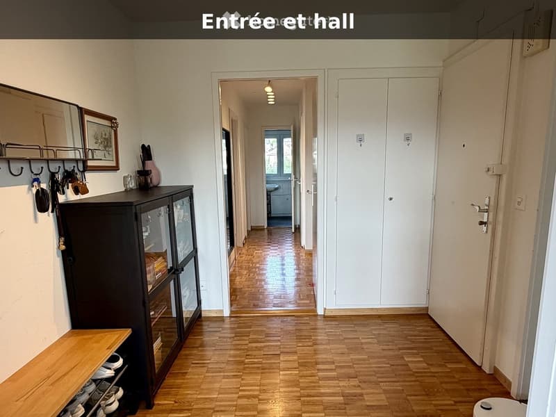 Entrance and hallway with wooden flooring, white walls, and a coat rack with keys and bags. A small wooden bench holds shoes, and a dark cabinet with glass doors is visible.