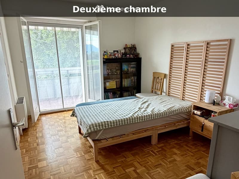 Cozy bedroom with wooden flooring, a bed with a checkered duvet, a nightstand, a chair, a radiator, and a bookcase with decorative items.