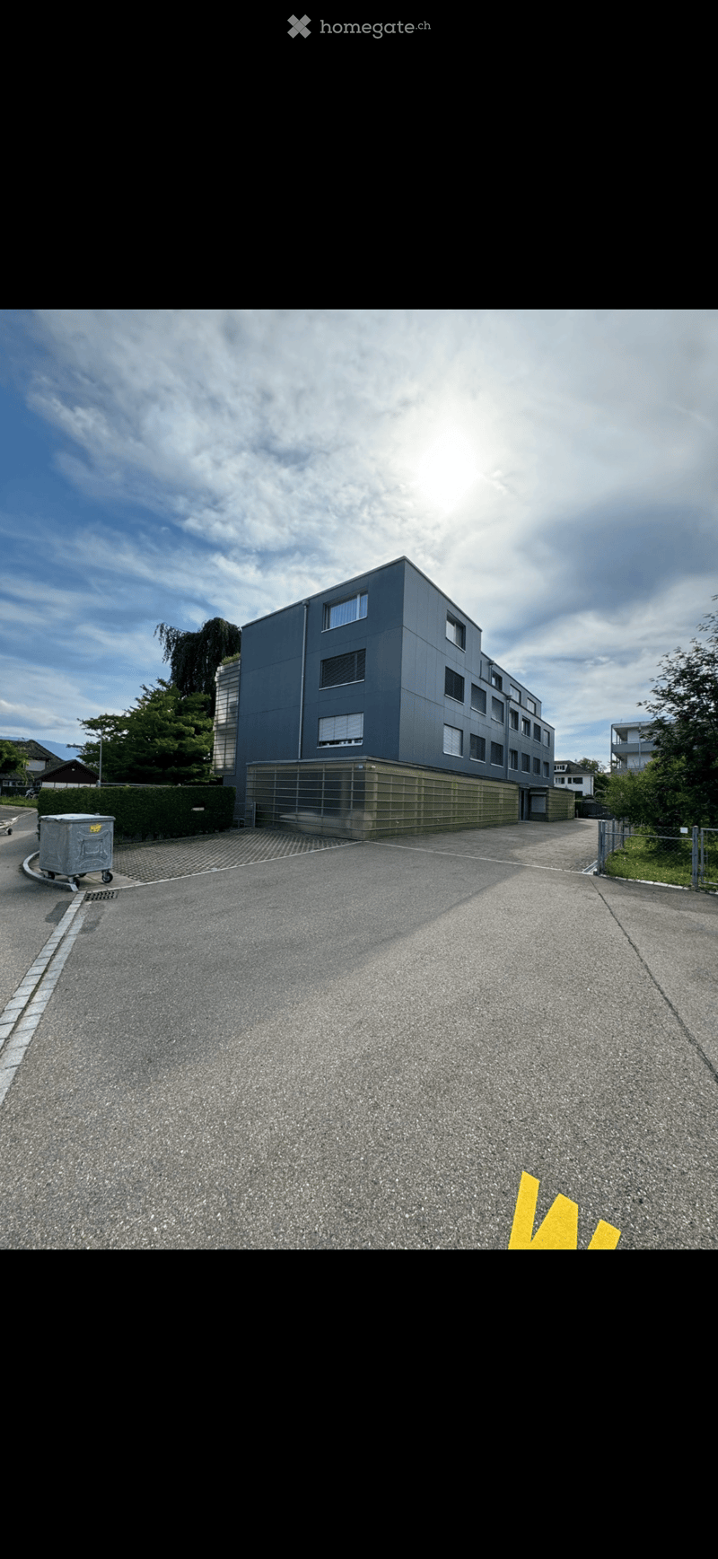 Modern multi-story building with grey facade and large windows, surrounded by greenery and a paved driveway.