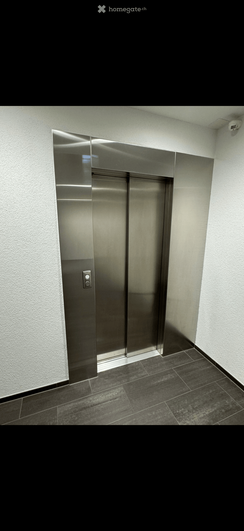 Modern hallway with dark tiled floor, light-colored walls, and a stainless steel elevator door with control panel.