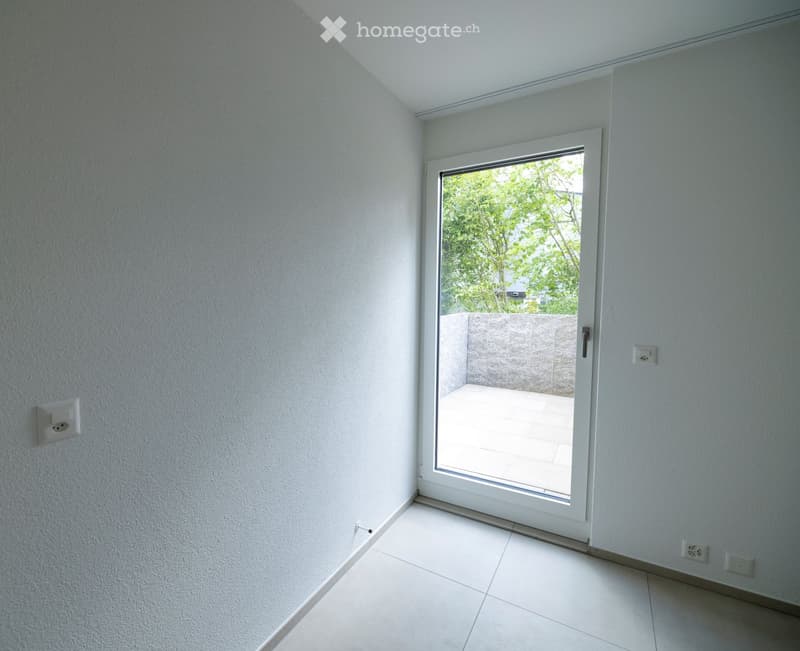 Empty room with white tiled floor, white walls, and a large sliding glass door leading to an outdoor area with a stone wall and greenery visible.
